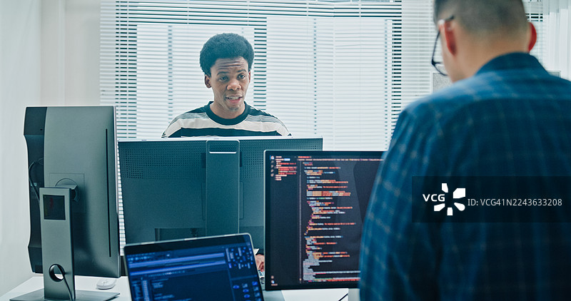 两位男性软件开发人员在办公室的办公桌前，通过多个屏幕分析数据、编写代码并工作。涉及人工智能和编程。图片素材