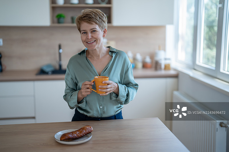 一位成熟的女士在厨房里享用早晨的咖啡和糕点，面带微笑图片素材