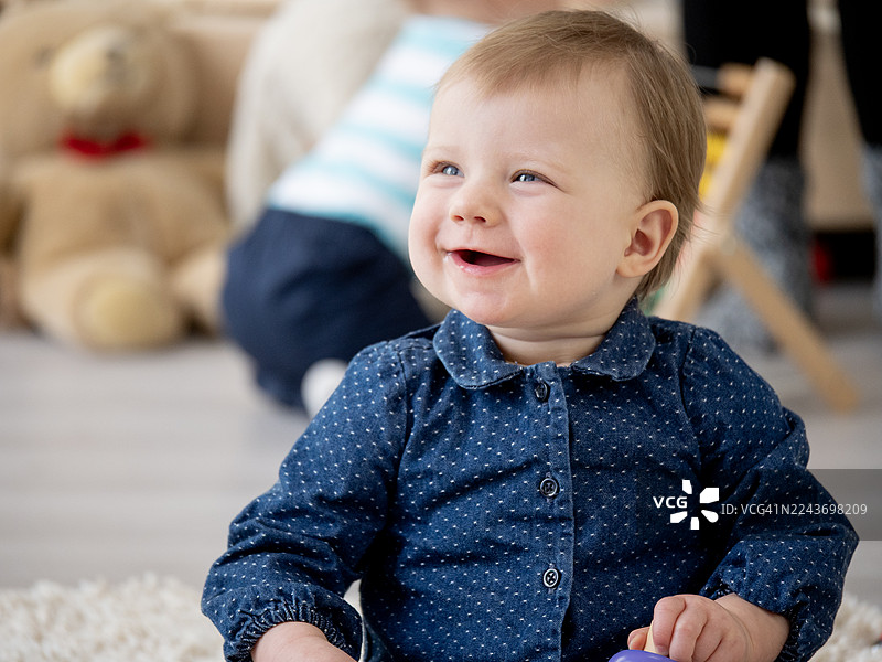 穿着牛仔衬衫的快乐宝宝在家中明亮的背景下微笑，周围有毛绒玩具，展现着玩耍的温馨瞬间图片素材
