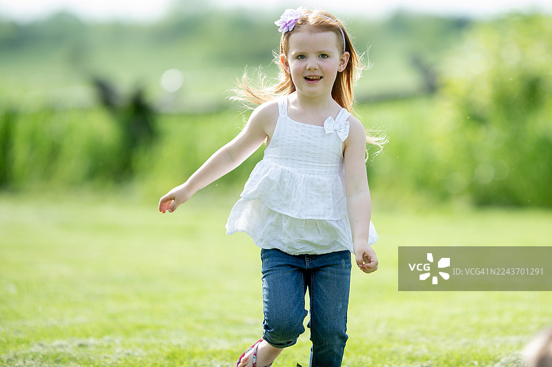 阳光明媚的户外，快乐的小女孩穿着白色上衣和牛仔裤，在绿色的田野里玩耍。图片素材
