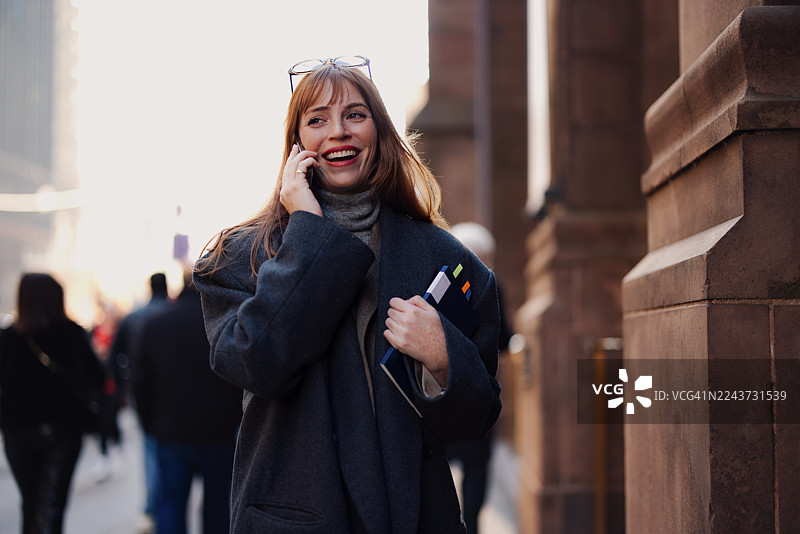 一位戴着眼镜、身穿深色外套的女士，在纽约的街道上微笑着打电话。图片素材