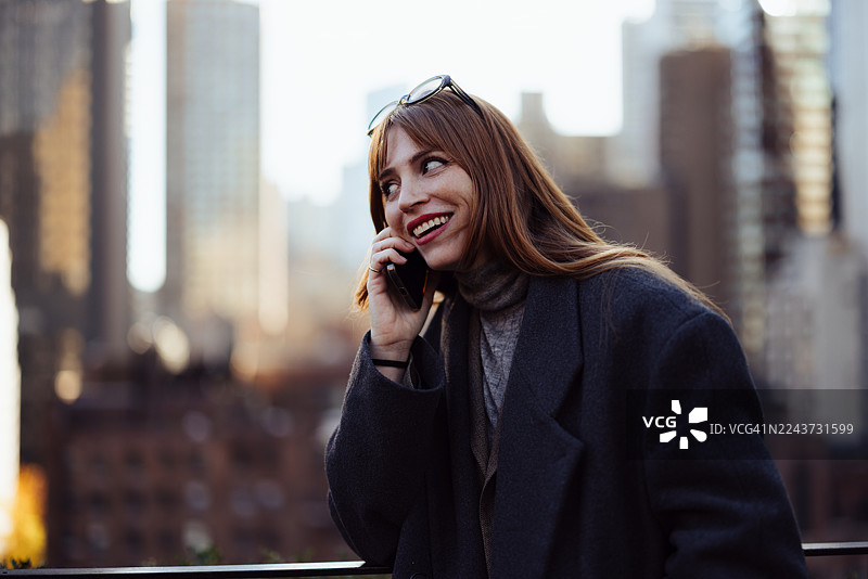 穿着深色外套的女士，手持电话，俯瞰着繁忙的纽约市屋顶天际线，脸上带着笑容图片素材