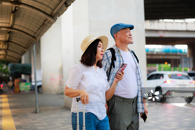 亚洲老年夫妇在度假旅行中，在街上等出租车时使用手机，展示了快乐的退休旅行者在户外使用科技来导航城市交通。图片素材