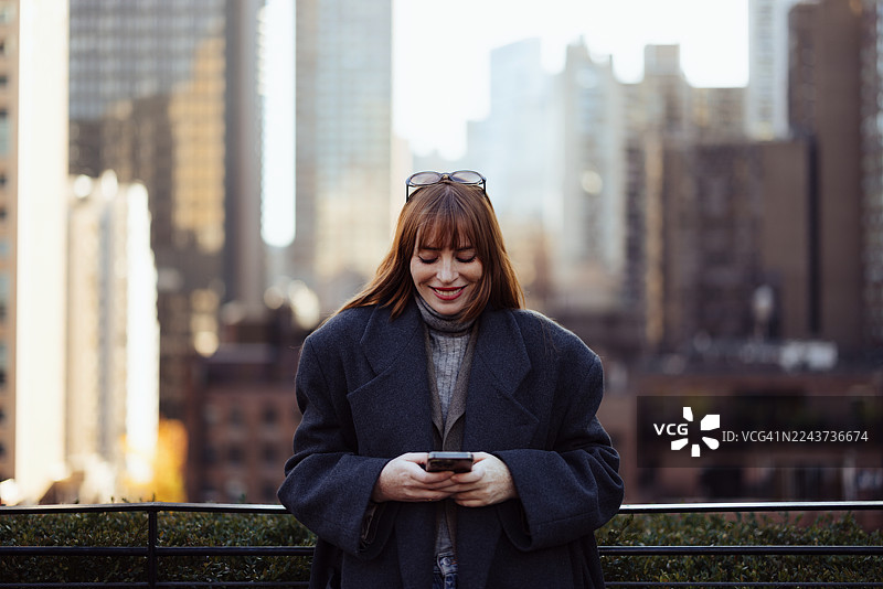 戴眼镜的女士身穿暖和的外套，在纽约市天际线背景下使用手机图片素材