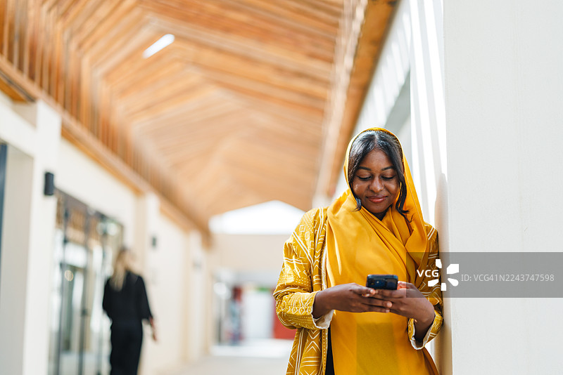 穿着黄色阿巴亚的时尚中年黑人女性正在查看智能手机图片素材