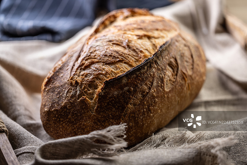 一个匠人手工制作的面包，拥有金黄酥脆的外皮。面包放在一张褶皱的浅色亚麻布毛巾上，毛巾置于一个乡村风格的深色木桌上。图片素材