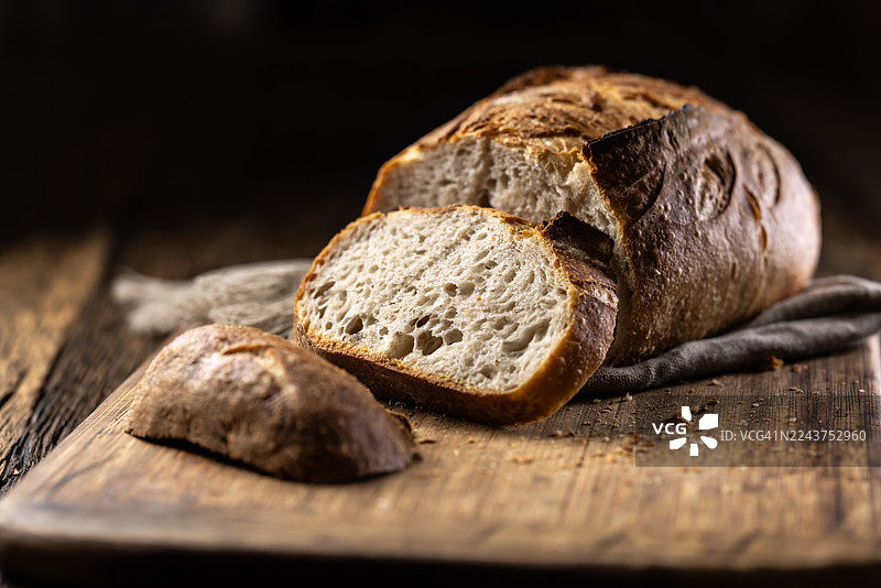 一个手工制作的酸面包，部分切开，放在深色、乡村风格的木桌上，旁边有一大一小两片。面包放在一块木质砧板上。图片素材