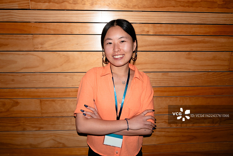 一位年轻女子自信地双臂交叉而立，身穿亮橙色衬衫，佩戴着工作识别证。图片素材