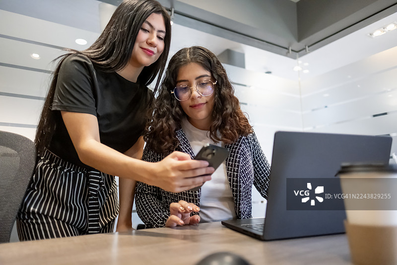 年轻的女高管们在现代化的办公室环境里，快乐而勤奋地使用笔记本电脑和手机工作。图片素材
