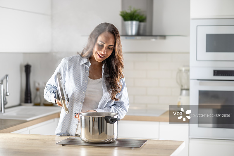 一位满意的家庭主妇，留着棕色长发，穿着浅蓝色条纹衬衫，正在明亮现代的厨房里打开不锈钢压力锅。锅盖和锅体冒着热气，表明食物已煮熟，锅盖很烫。图片素材