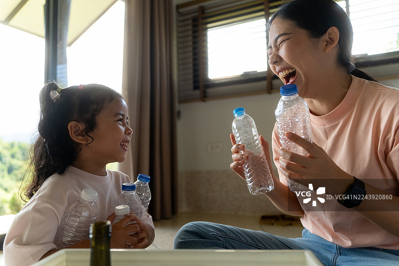 快乐的年轻亚洲母亲和她的小女儿在家中开心地笑着，进行着回收塑料和垃圾分类的活动，这是关于废物管理和全球环境生态的考量。图片素材