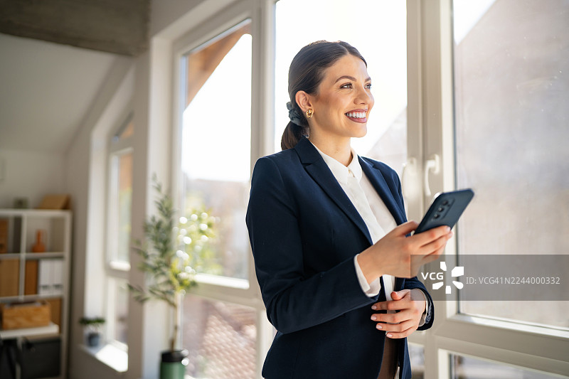 年轻的职业女性在办公室，手持智能手机，望向窗外图片素材