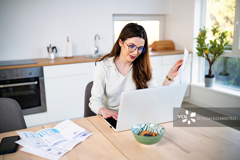 年轻女子在家使用笔记本电脑工作，手中拿着财务报表图片素材