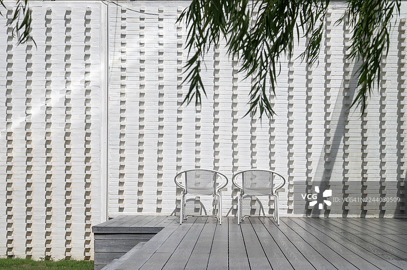 两张白色椅子摆放在木质露台上，椅子面对面放置。露台由木材制成，后面是一堵白色砖墙。图片素材
