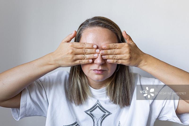 一位女士双手遮住眼睛，做出俏皮的“非礼勿视”姿势，她身穿白色T恤，背景是中性色调图片素材