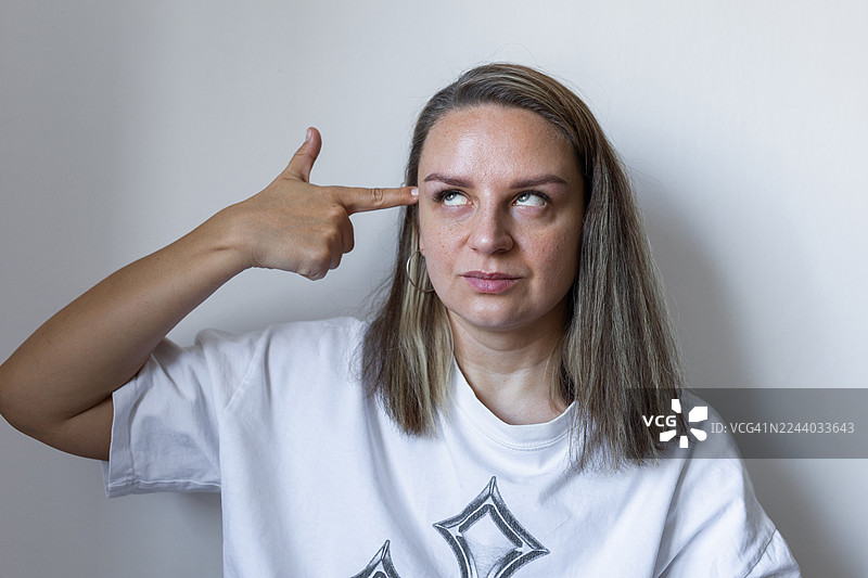 一位穿着休闲白色T恤的女性，做出用手指指向头部的沉思姿势图片素材