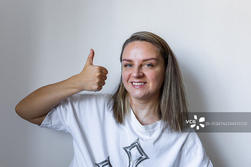 自信的女性身穿休闲白色T恤，竖起大拇指，面带微笑的肖像照图片素材