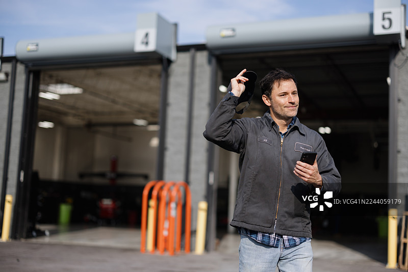 汽车修理店里，一位技师在打招呼，同时手中拿着一部智能手机。图片素材