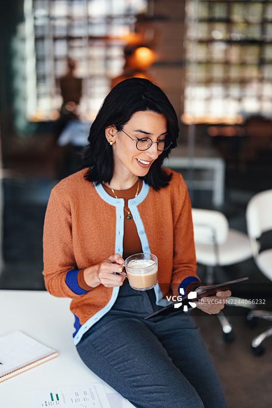 自信的女性在现代办公室环境中，一边喝咖啡一边使用平板电脑工作图片素材