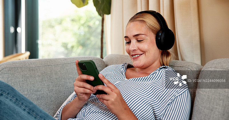 快乐的女子戴着耳机，坐在沙发上，用手机播放音乐、在线视频，享受周末时光。她面带微笑，在家里用智能手机听播客、音频播放列表或网络剧。图片素材