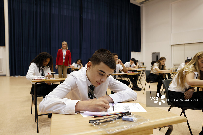 一名中学生在一间宽敞的学校礼堂里参加考试，在一个安静的学术环境中专注于试卷。图片素材