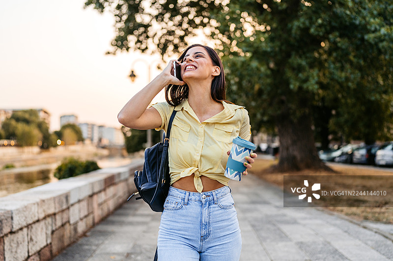 年轻女子在码头边边走边喝着外带咖啡，并用手机通话。图片素材