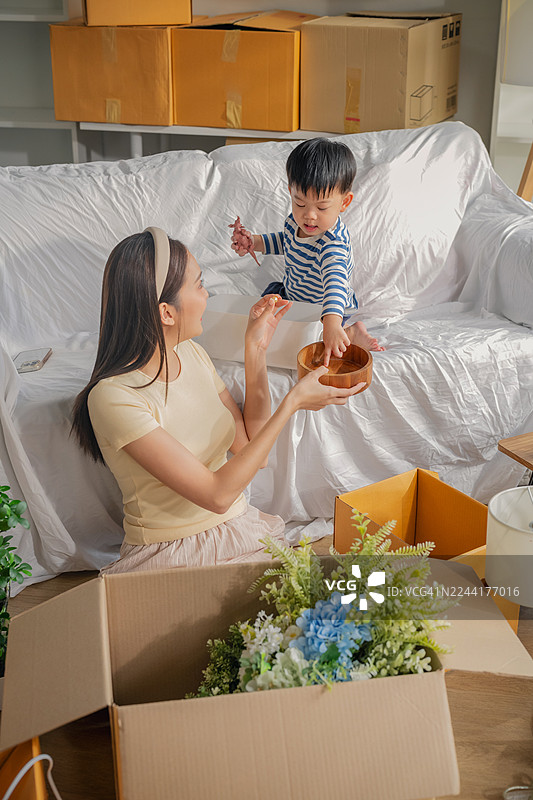 亚洲母亲在新家客厅拆箱，儿子坐在沙发上玩玩具，一家人搬家、翻新和新房采光图片素材