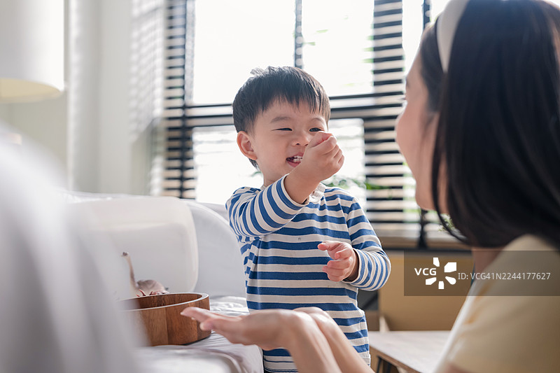 亚裔家庭温馨互动，母亲和儿子坐在沙发上放松玩耍，脸上洋溢着欢笑，充满愉悦的氛围。背景是搬家用的纸箱，暗示着搬迁。她非常爱她的儿子。图片素材
