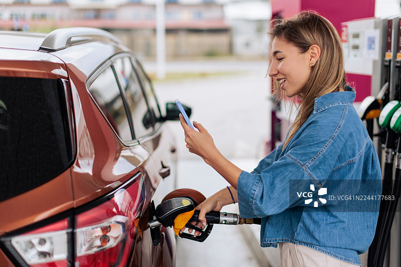 微笑着的女士在给汽车加油时使用手机图片素材
