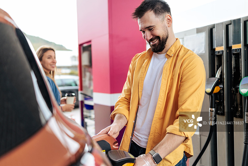 一对快乐的夫妇在加油站，手里拿着外带咖啡，为汽车加油图片素材