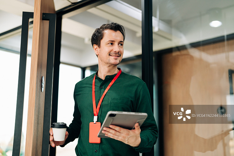 专业男士站在玻璃门旁，手中拿着平板电脑和外带咖啡图片素材