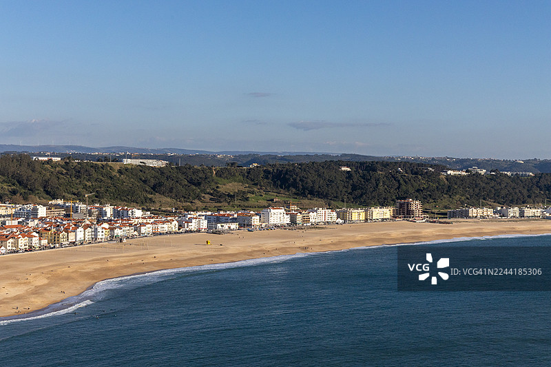 郁郁葱葱的山坡旁坐落着一个充满活力的海滨小镇的全景，深邃的蓝色海洋在晴朗的天空下绵延开去图片素材