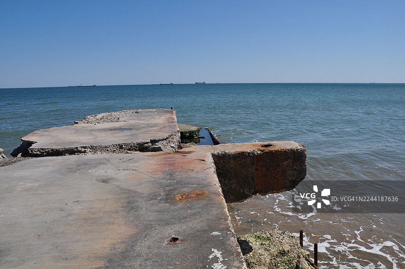 晴朗的天空下，饱经风霜的混凝土码头伸向宁静的蓝色大海，远处的地平线上有船只，中国北戴河图片素材