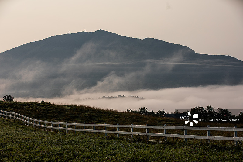 远眺被薄雾和云层半遮半掩的山景，前景是白色栅栏和绿草，美国佛蒙特州东伯克。图片素材