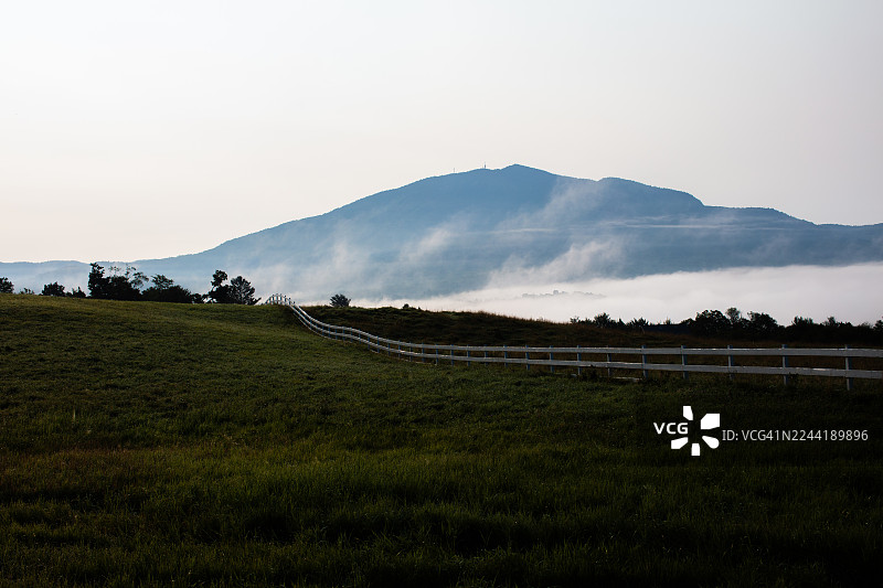 晨雾弥漫的乡村风景，白色栅栏蜿蜒穿过绿色的田野，延伸向笼罩在薄雾中的远山。美国，东伯克。图片素材