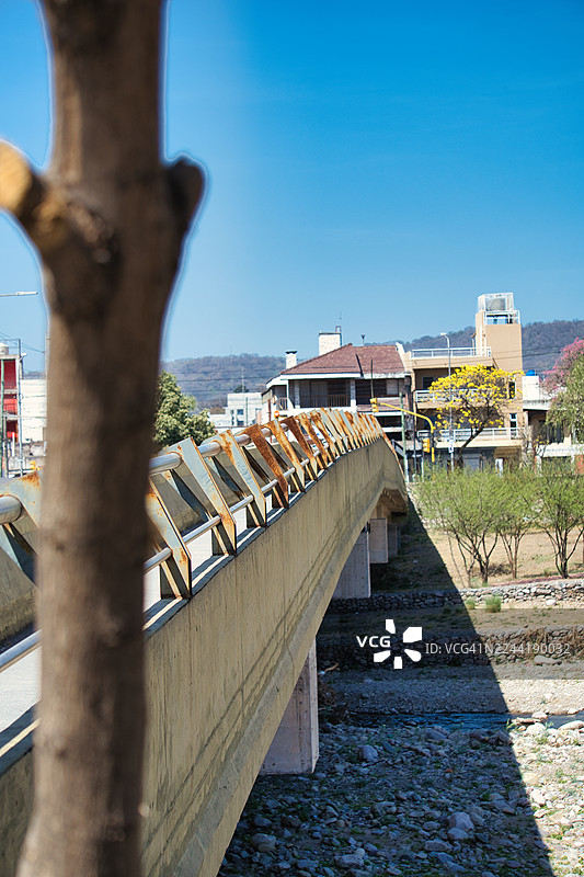 一座行人天桥，锈迹斑斑的栏杆横跨干涸的河床，树干作为前景，背景是建筑和山脉，天空湛蓝，阿根廷胡胡伊省图片素材