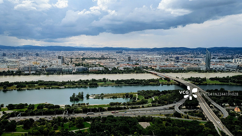 阴云密布的天空下，高角度俯瞰城市，可见河流和高速公路立交桥，展示着城市发展与自然元素的融合，奥地利维也纳图片素材