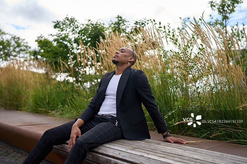 穿着时尚黑色西装外套的休闲男子，在自然城市公园的长椅上向后倚靠，周围环绕着高高的草丛和绿植。图片素材