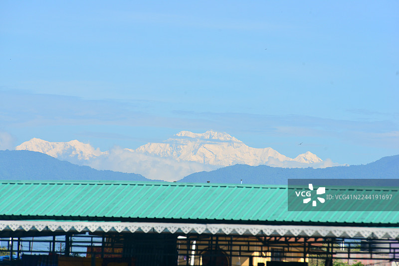 在晴朗的蓝天下，雄伟的雪山高耸于远处的绿色屋顶之上，唤起了宁静与自然之美之感。图片素材