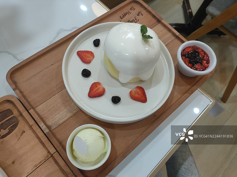 俯视视角拍摄的日式舒芙蕾松饼，蓬松柔软，配有奶油、草莓和蓝莓，摆放在白色盘子上，旁边是木质托盘上的莓果酱和冰淇淋图片素材