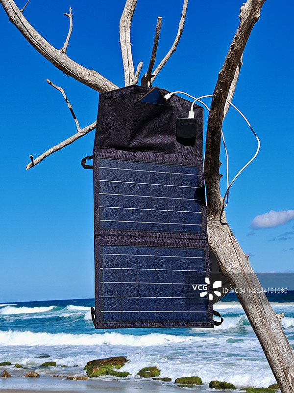 阳光明媚的海滩上，便携式太阳能电池板充电器挂在光秃秃的树枝上，为智能手机充电，背景是保加利亚的海景。图片素材