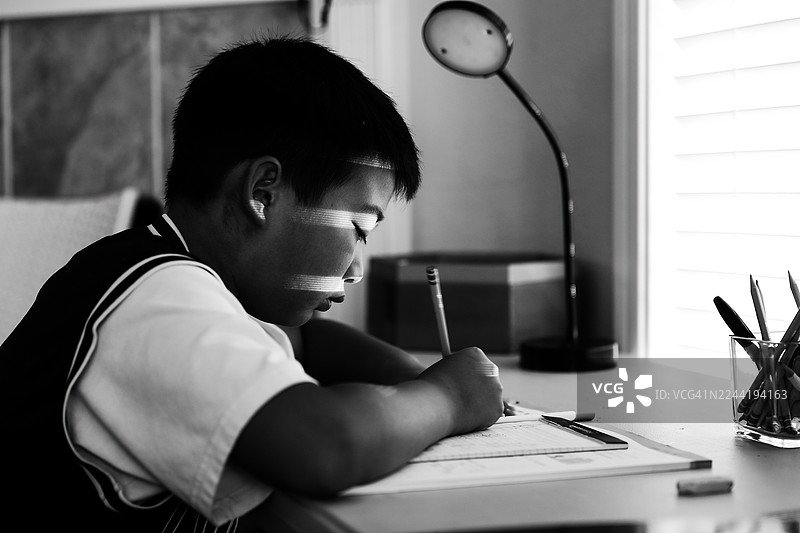 一个小男孩正专注地在书桌前写字，脸上掠过一道道光影。图片素材