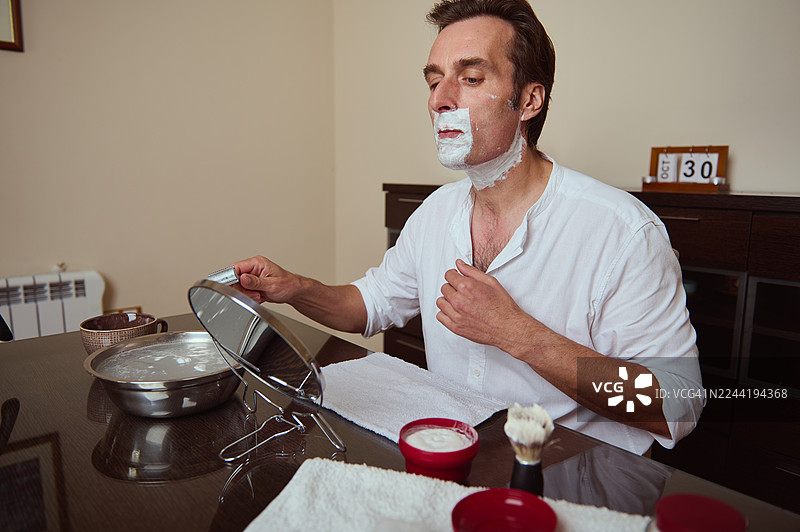 男人在家刮胡子，脸上涂着剃须泡沫，背景是镜子和日历显示“今天”图片素材