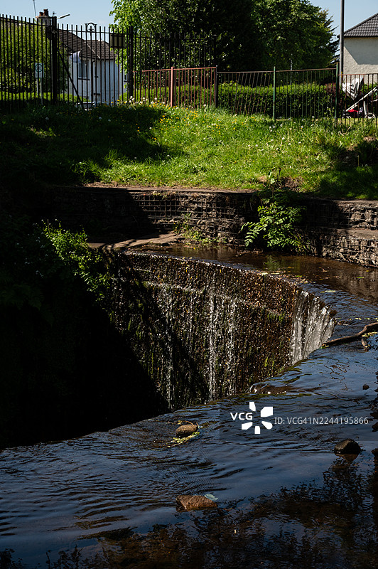 苏格兰达尔穆尔一处河流及其小型瀑布的风景，周围环绕着郁郁葱葱的绿植和远处的建筑，天空晴朗。图片素材