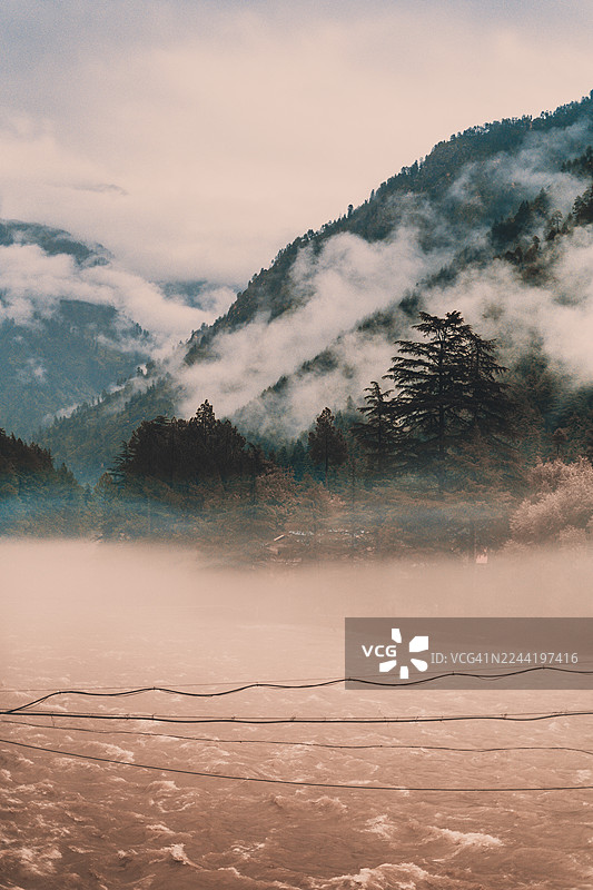 薄雾笼罩的山景，一条河流淌而过，茂密的常绿森林在云层密布的天空下，印度马尼卡兰，卡索尔山丘图片素材