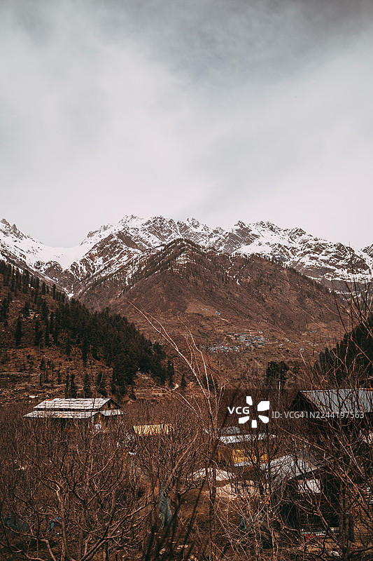 印度的马纳利，阴天笼罩下，白雪皑皑的山峰耸立在山谷之上，山谷中散布着稀疏的村舍建筑和光秃秃的树木，景色壮丽。图片素材