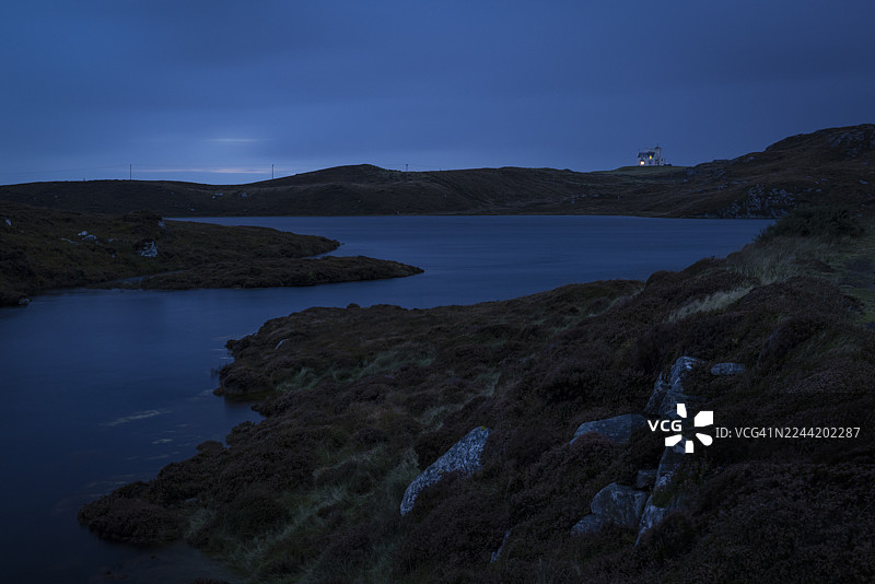 苏格兰刘易斯和哈里斯岛，宁静的湖泊倒映着渐暗的天空，石楠覆盖的山丘与水边相接，远处的房屋散发着柔和的光芒。图片素材