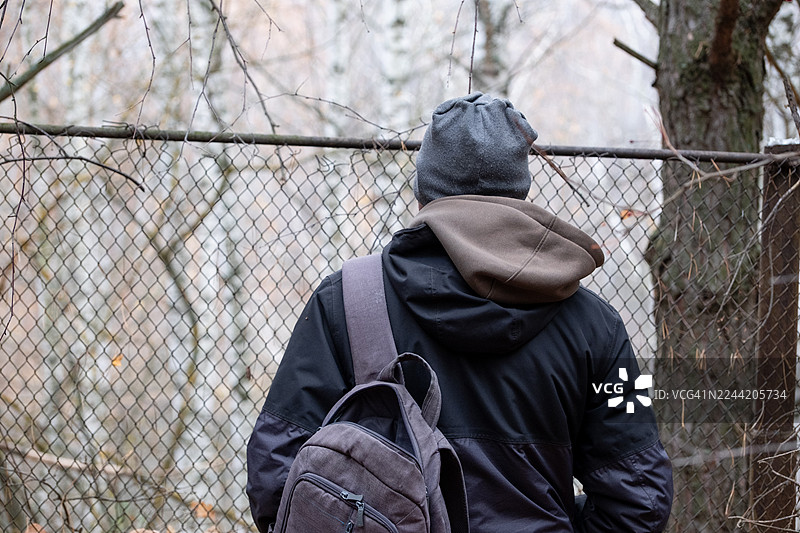 一名穿着夹克、背着背包的男子站在森林中的边境口岸围栏前。图片素材