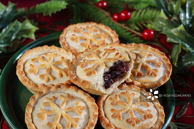 特写图片，绿色盘子里装着一堆肉馅饼，酥皮圣诞甜点，少了一口，露出甜肉馅，塑料杉树枝，绿色冬青叶带红浆果，斑驳的红色背景，前景聚焦图片素材