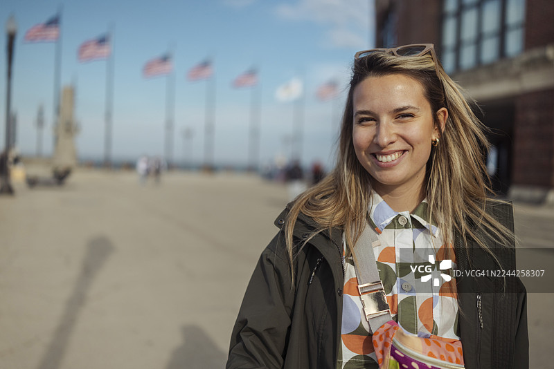 一位年轻女子面带微笑，身处芝加哥的城市景观之中，背景有美国国旗，天气晴朗明媚。图片素材